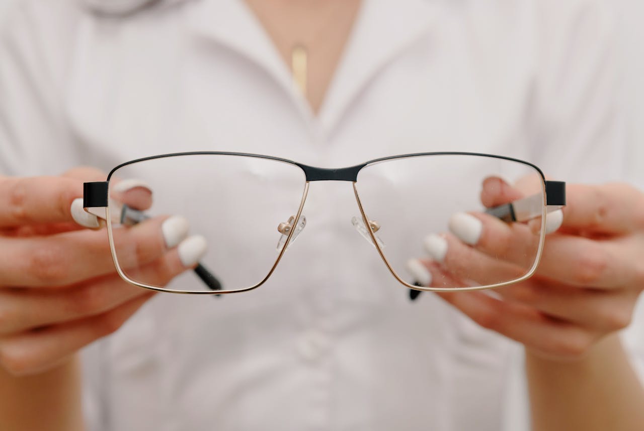 Crop anonymous female with manicured hands wearing white robe and showing classic eyeglasses in light modern optometrist clinic