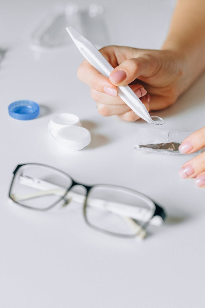 Closeup of a hand using a lens applicator with glasses and a contact lens case on a table.