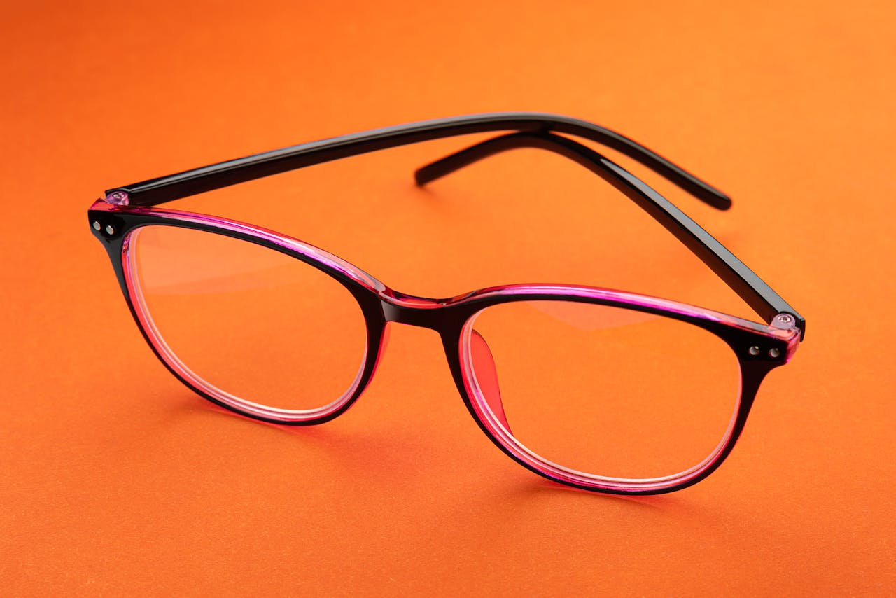 programs-03 Close-up of pink eyeglasses resting on a bright orange surface. Perfect for eyewear fashion concepts.